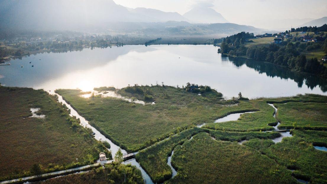 Luftaufnahme eines ruhigen Alpensees mit Feuchtgebieten und Spazierwegen bei Sonnenuntergang